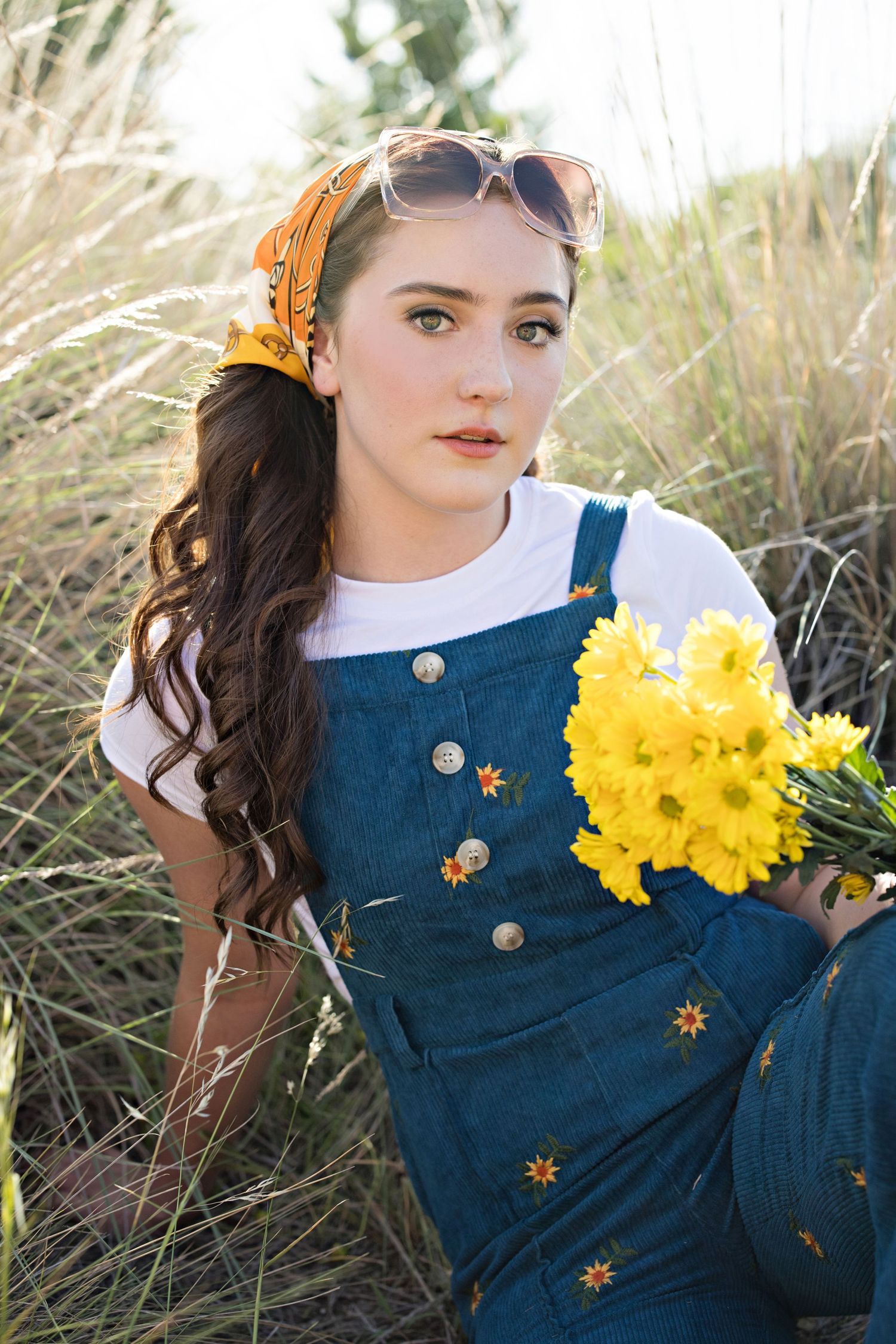 A person wearing denim overalls with embroidered flowers sits in tall grass holding bright yellow daffodils.