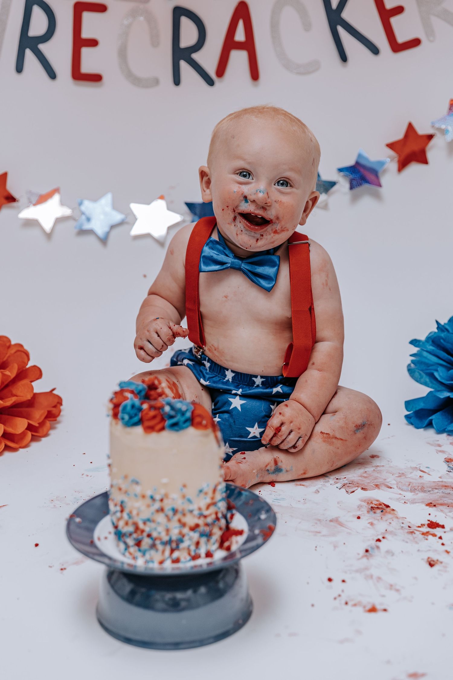 A baby in patriotic attire smiles while sitting next to a smashed cake during a photo session.