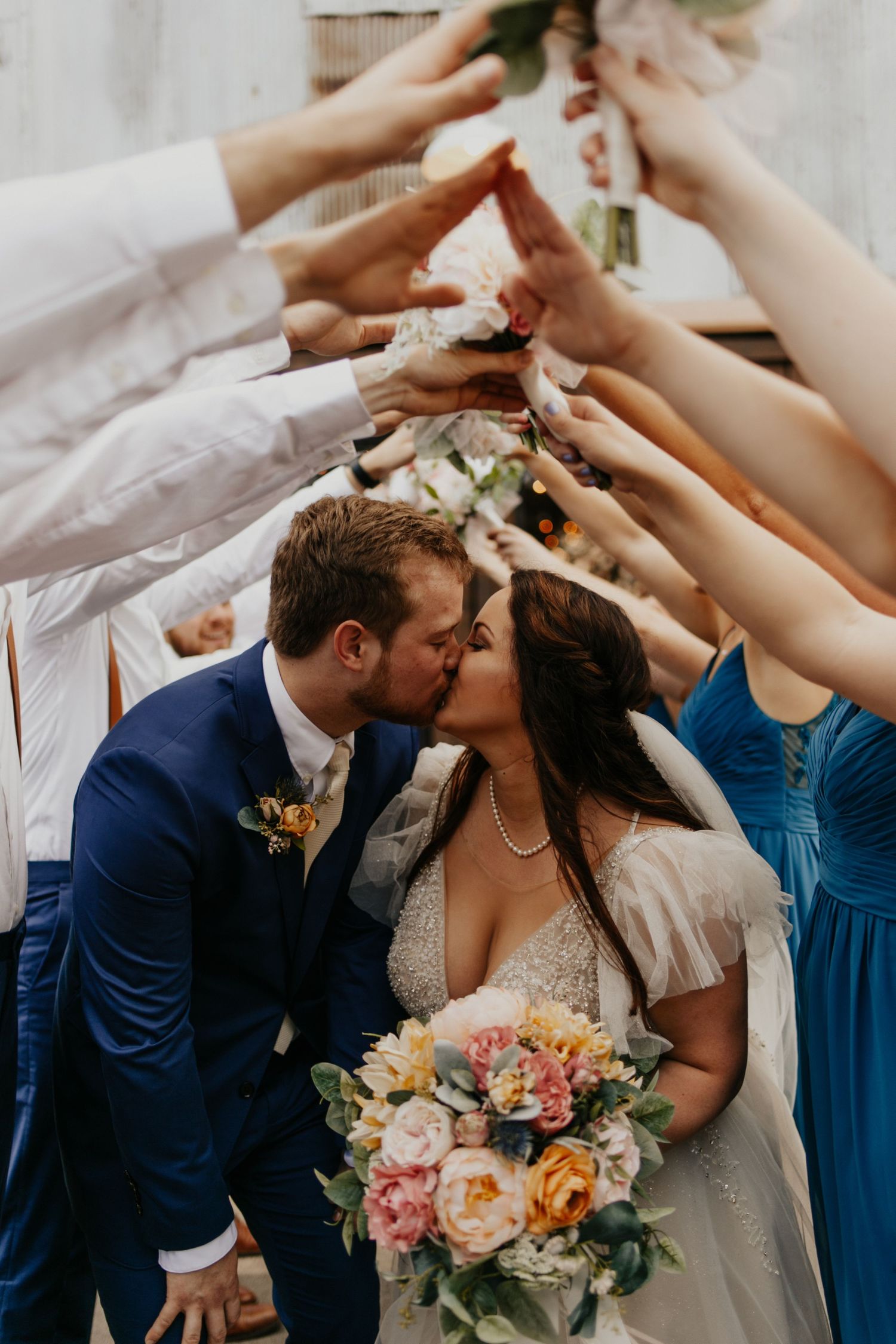 Newlyweds kiss under an arch of raised hands while holding a colorful bouquet at their wedding celebration.