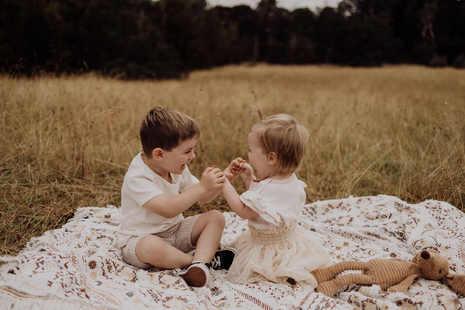 Two young children sit together on a white blanket in a golden field playing and laughing.