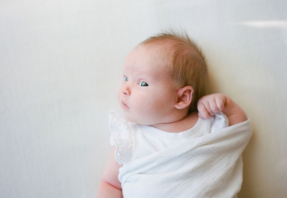 infant on white blanket wrapped in neutral swaddle looking curiously with head tilt at camera
