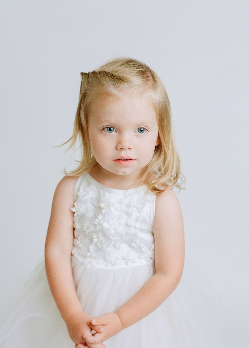 toddler blonde girl with blue eyes facing camera wearing white delicate dress with dainty white flowers