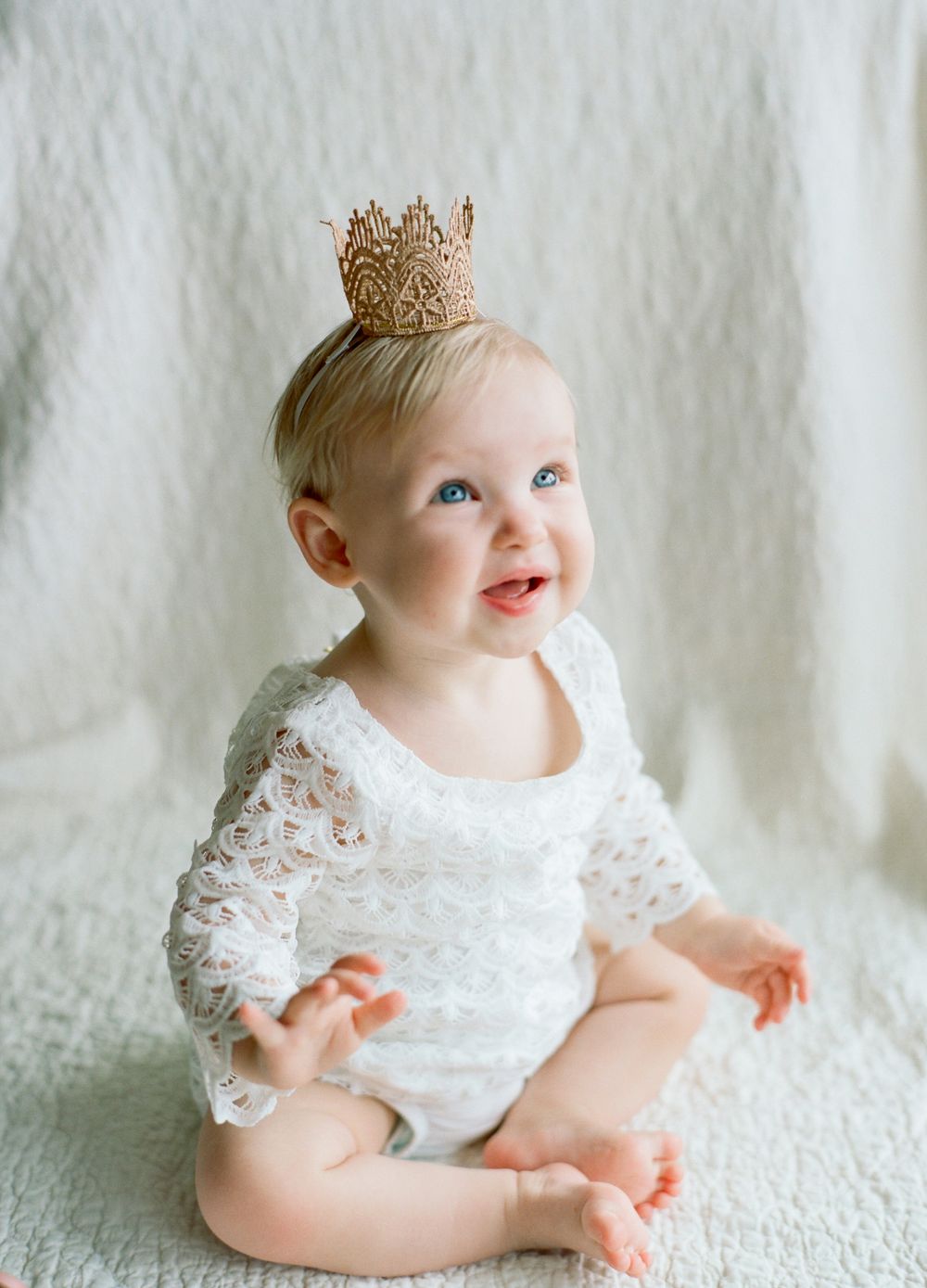 one year old  blonde baby girl wearing lace scalloped dress and a delicate gold crown in front of a soft white backdrop