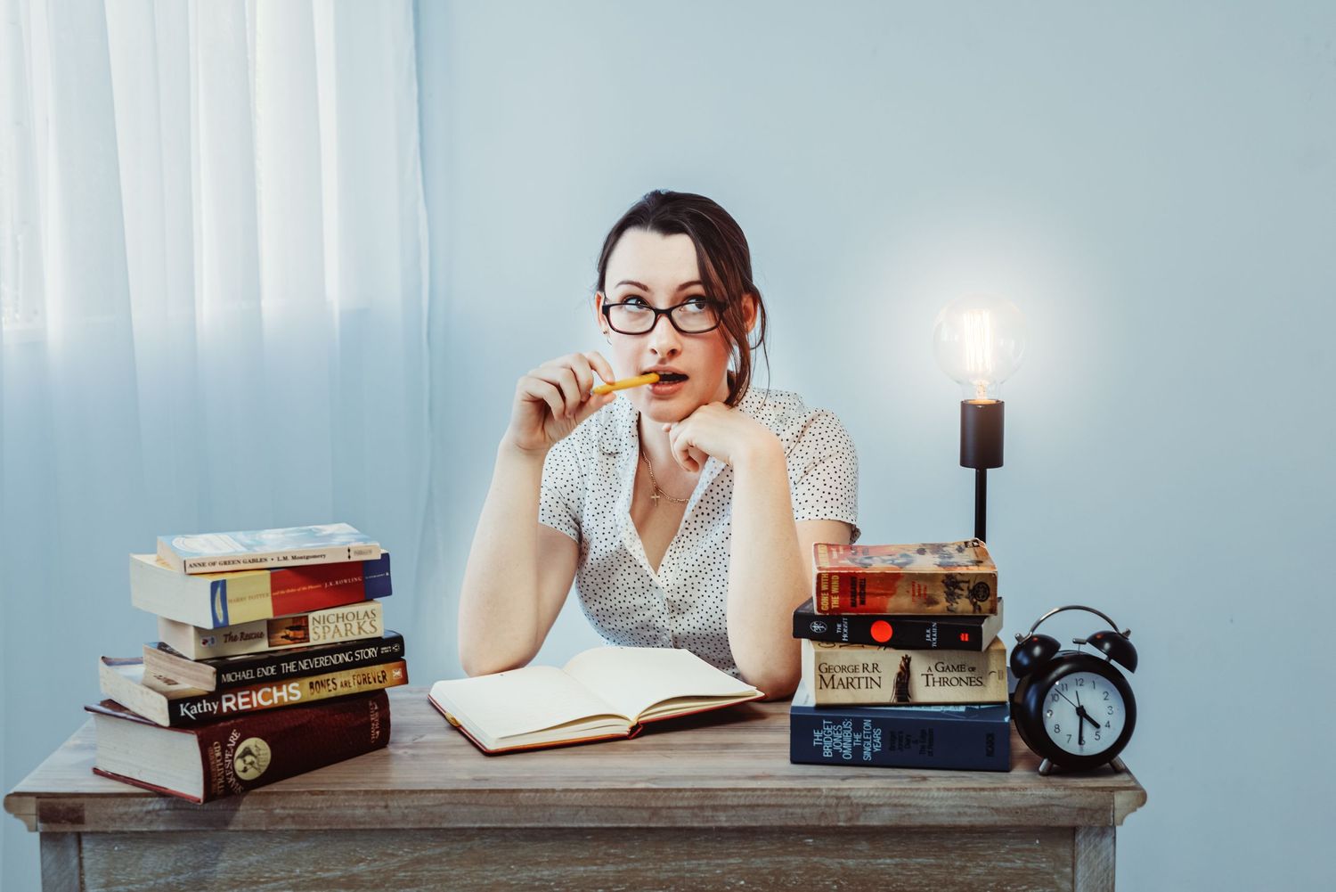 A study setup with stacked books, candle, and alarm clock on a desk with someone wearing glasses looking thoughtful.