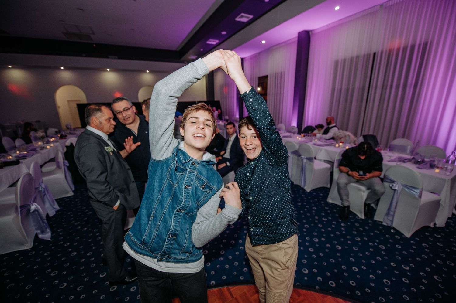 Wedding guests dance and celebrate with raised arms under purple venue lighting.