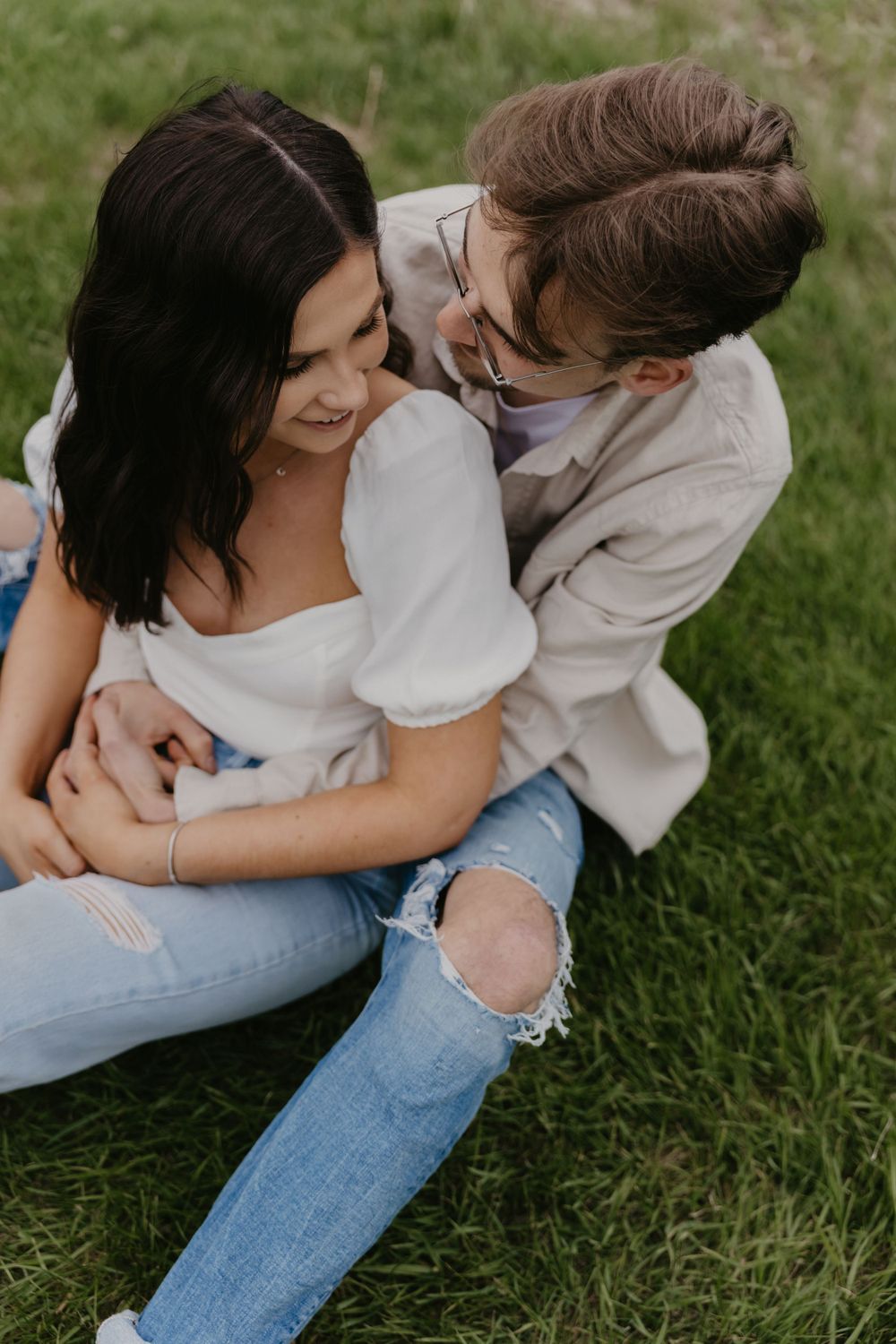 A couple shares laughter while sitting close together on grass during a sunny day.