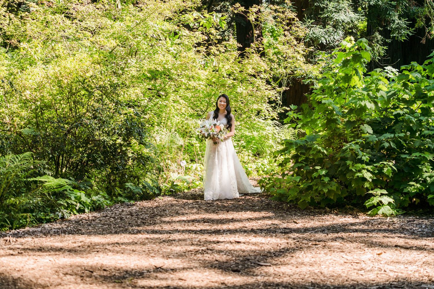A bride in a white gown stands on a sunlit forest path holding a bouquet.