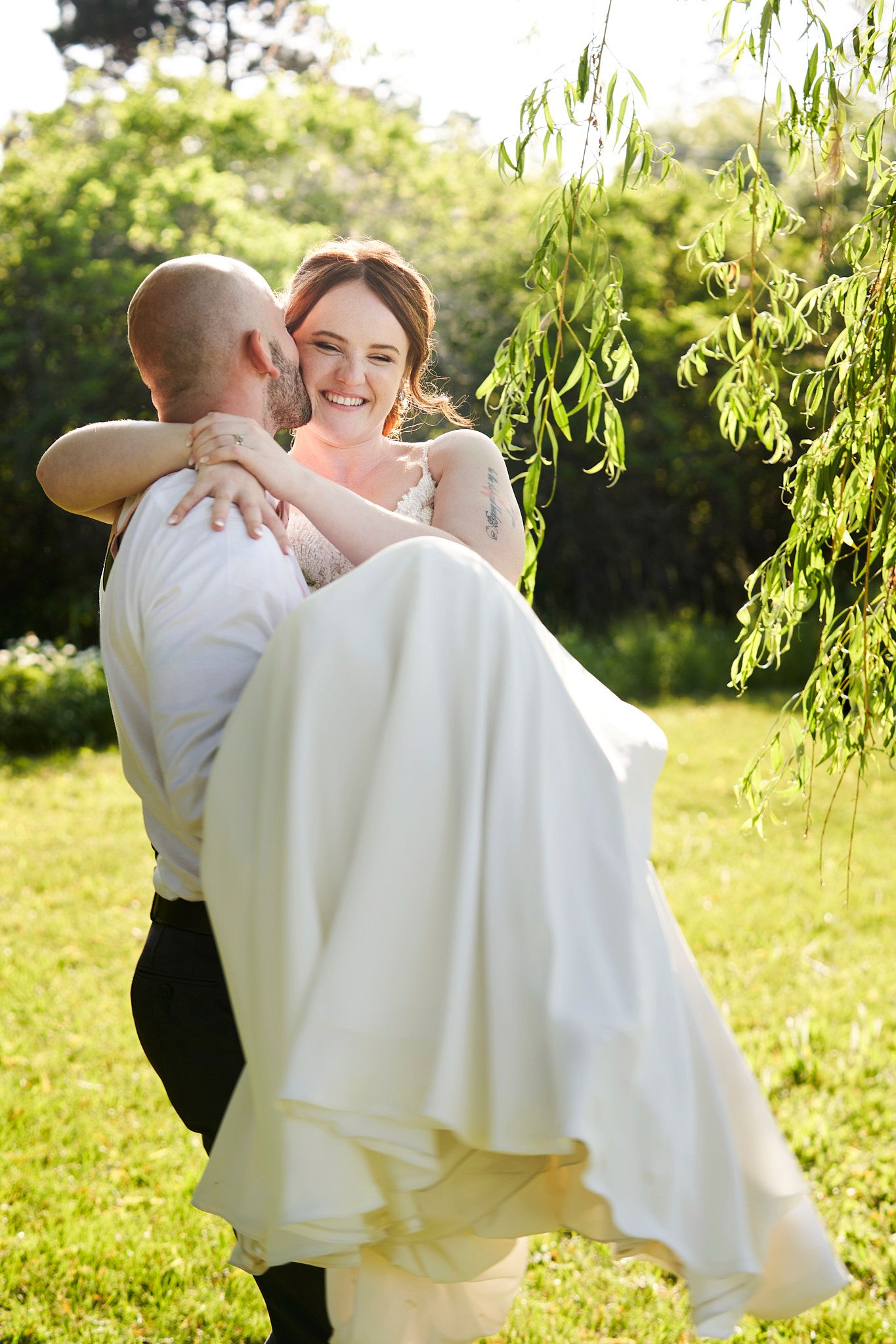 A joyful wedding couple in a garden setting with willow trees with the groom carrying his bride.