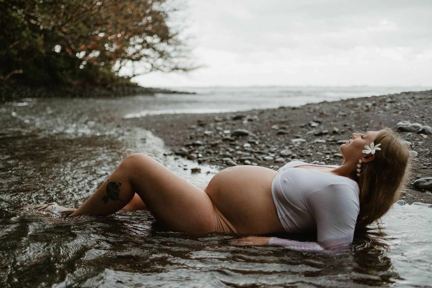 Maternity photo on rocky beach shore with person in white bodysuit at sunset.