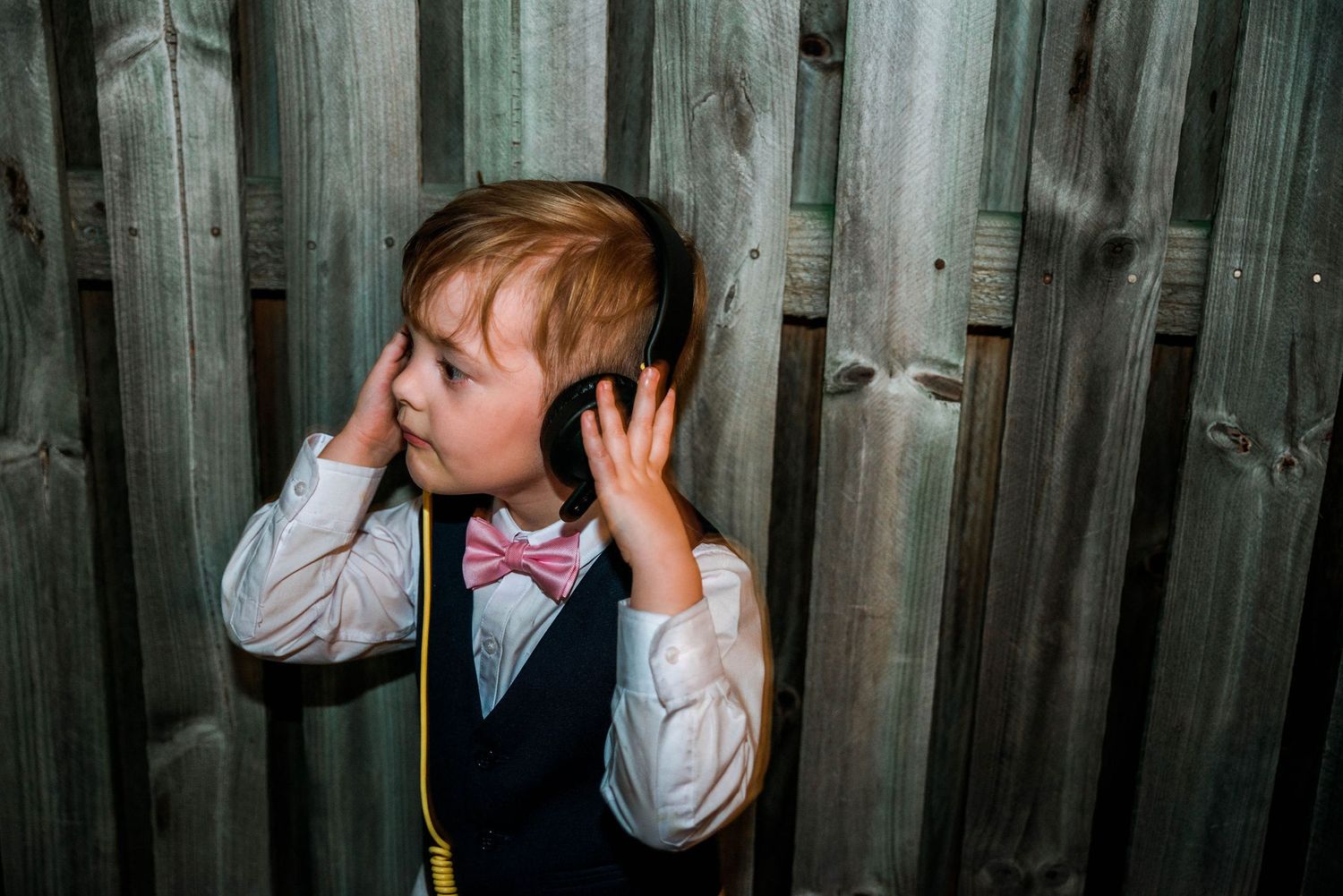 Young child in formal wear listening through headphones against wooden fence.