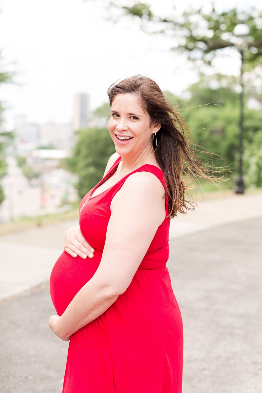 A pregnant woman in a bright red dress smiles while standing on a path with a city skyline behind her.