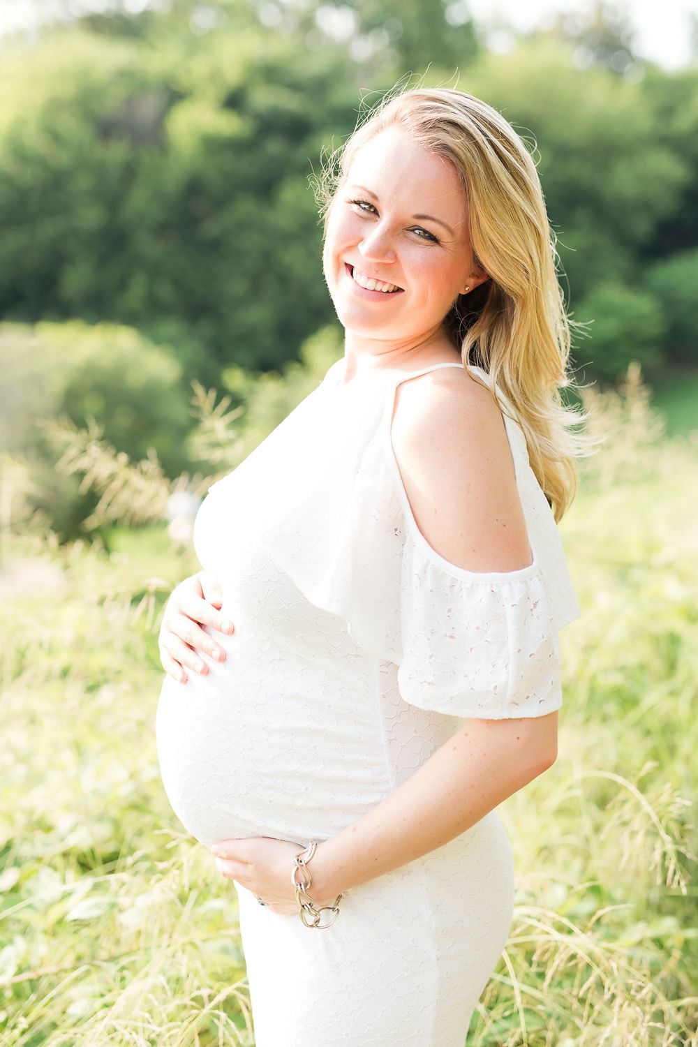 A pregnant woman in a white cold-shoulder dress poses in a sunny field.
