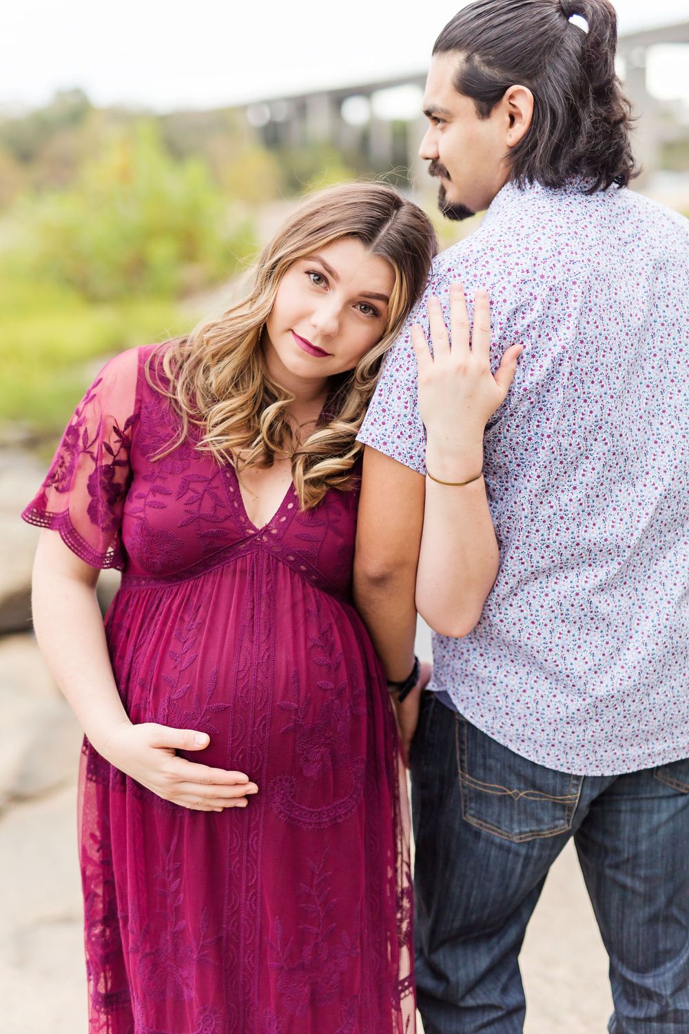 Couple embraces during outdoor maternity photo session near bridge.