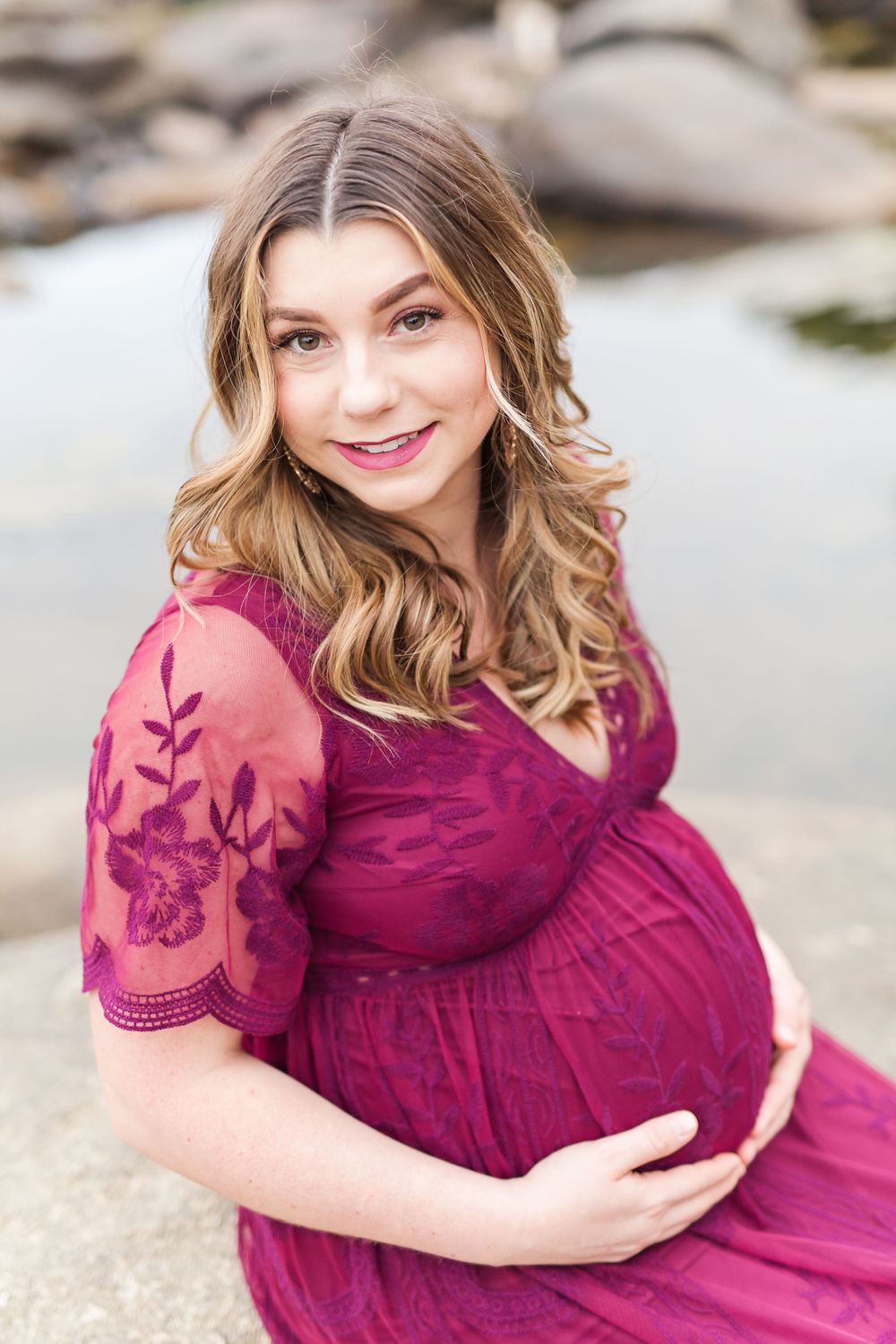 Person in burgundy lace dress poses during outdoor maternity photoshoot.