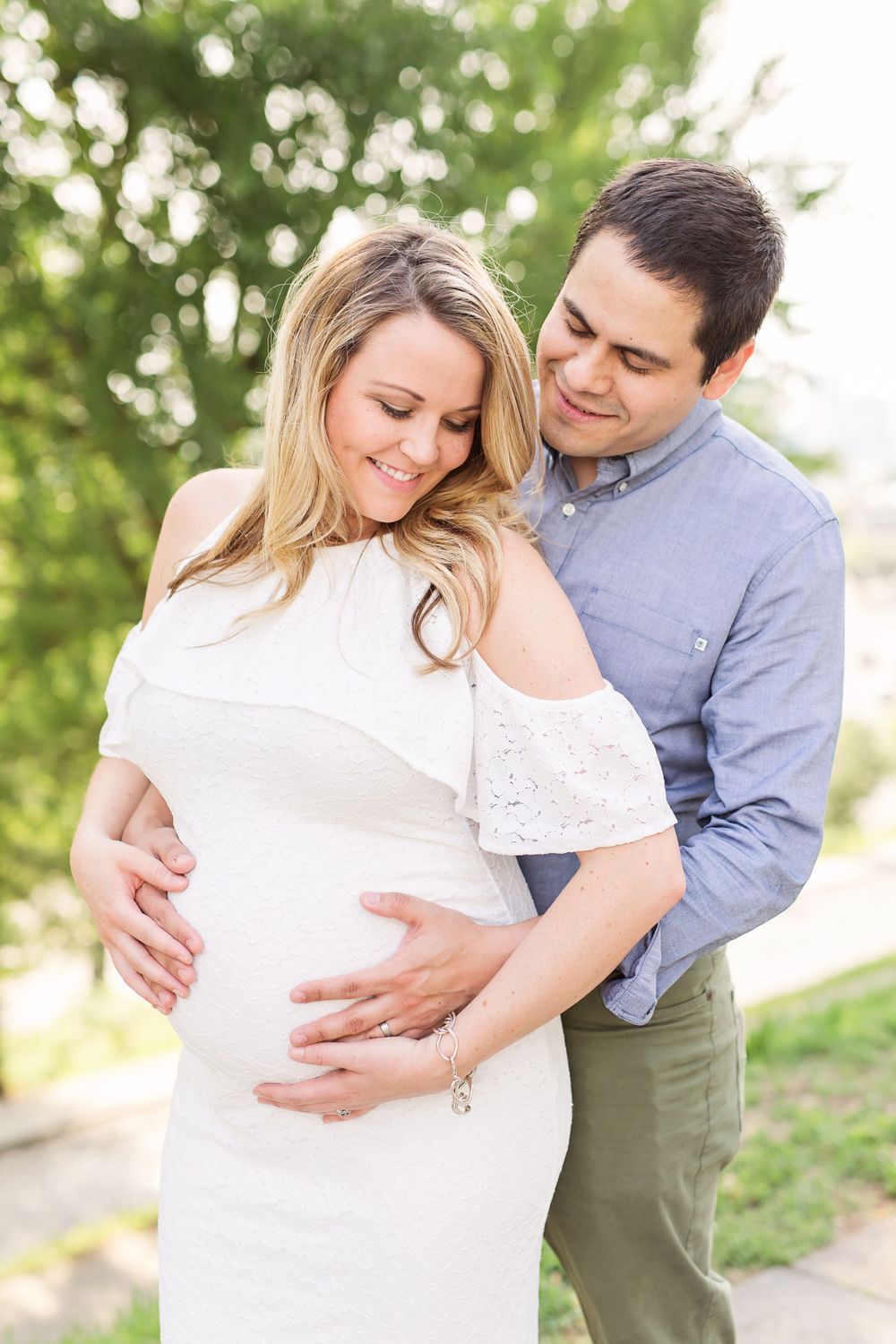 A maternity photo showing a couple embracing outdoors in a sunlit natural setting.