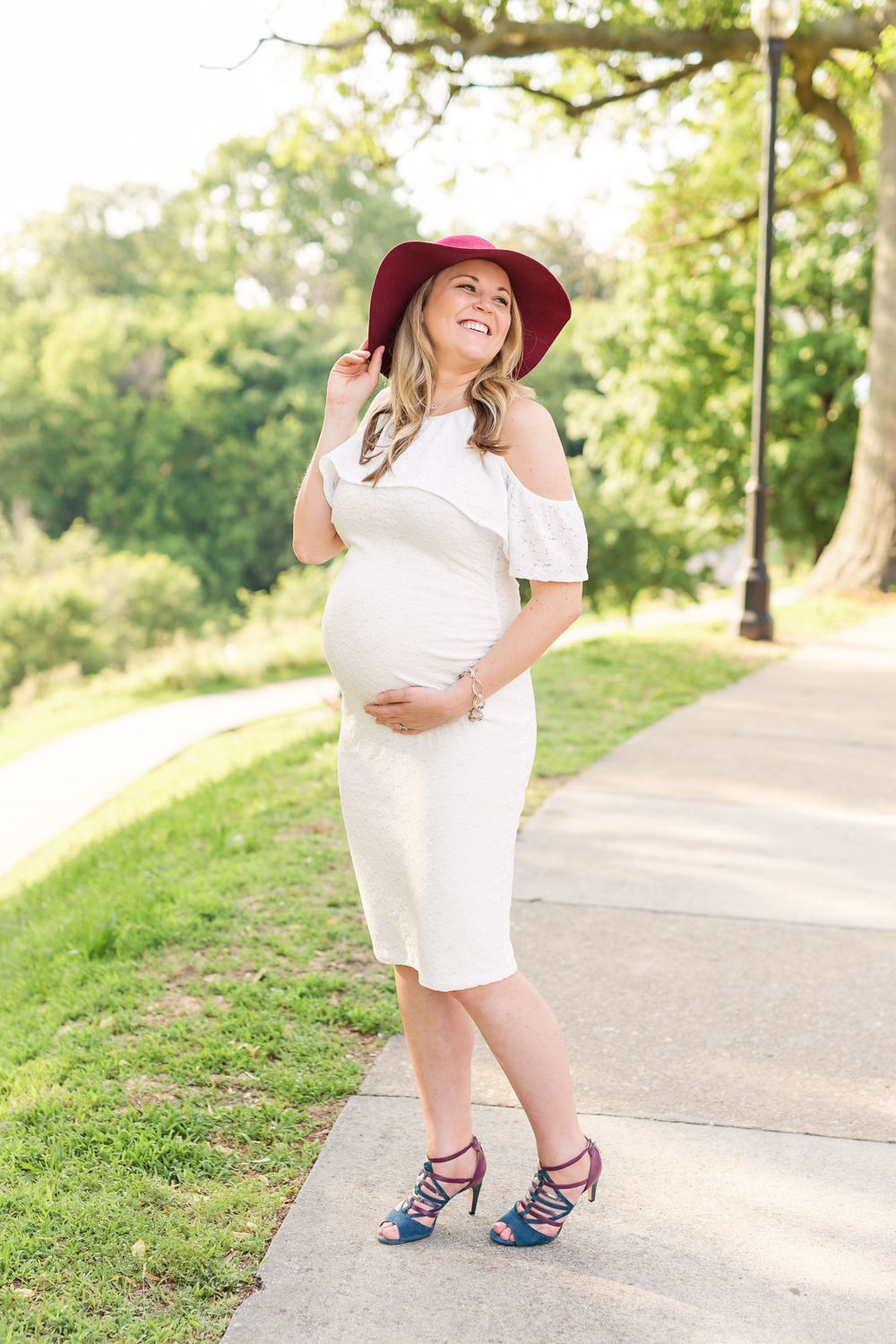 A pregnant woman in a white dress and burgundy hat poses on a pathway surrounded by greenery.