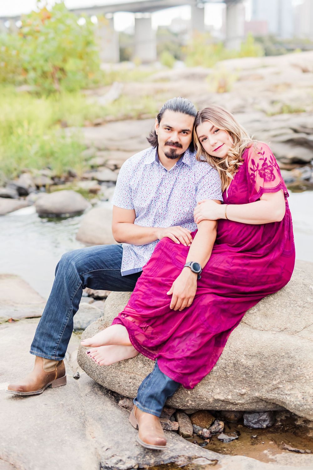A couple sits together on rocky terrain wearing casual attire with a burgundy dress and polka dot shirt.