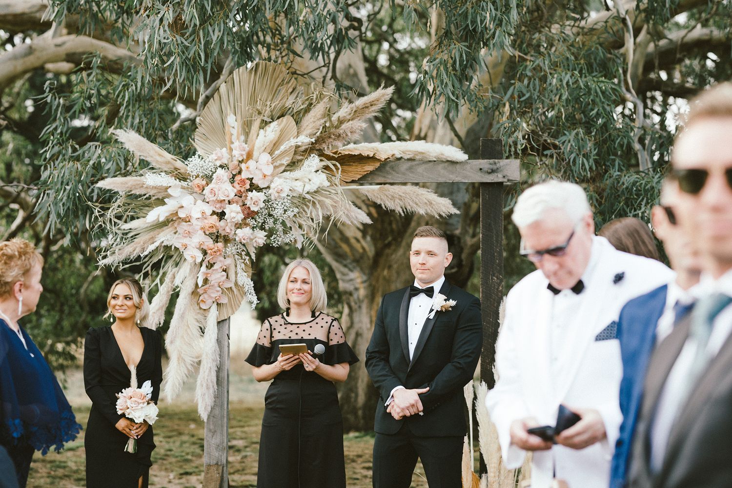 Wedding ceremony under eucalyptus trees with dried palm floral arch and guests seated on wooden chairs.