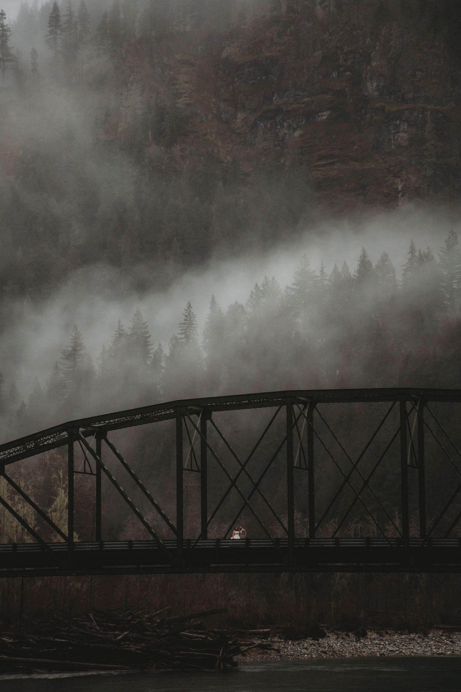 A bride and groom stand on a bridge under a giant mountain side at their mountain elopement.