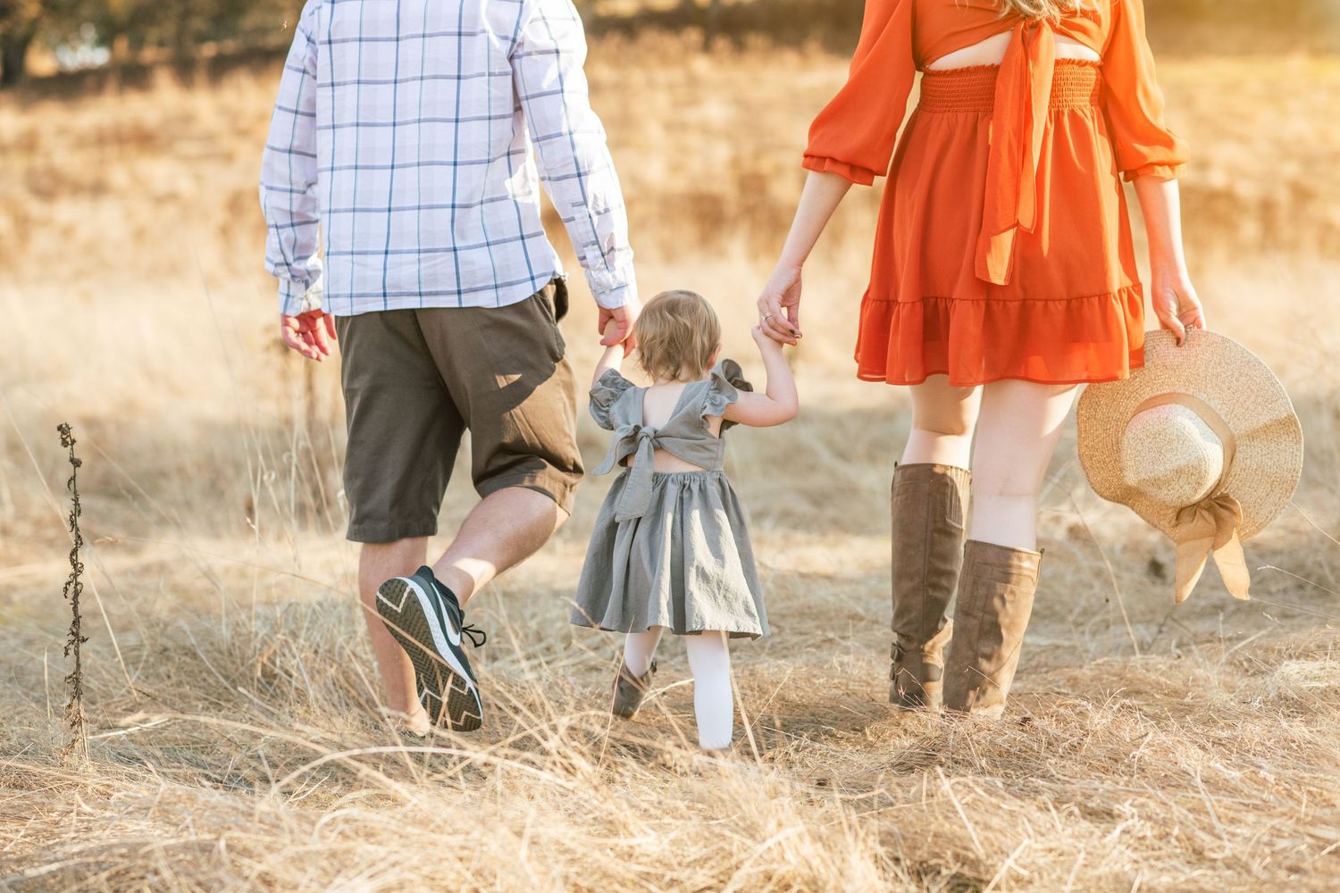 dad and mom holding young kids hand walking away from camera in a field.