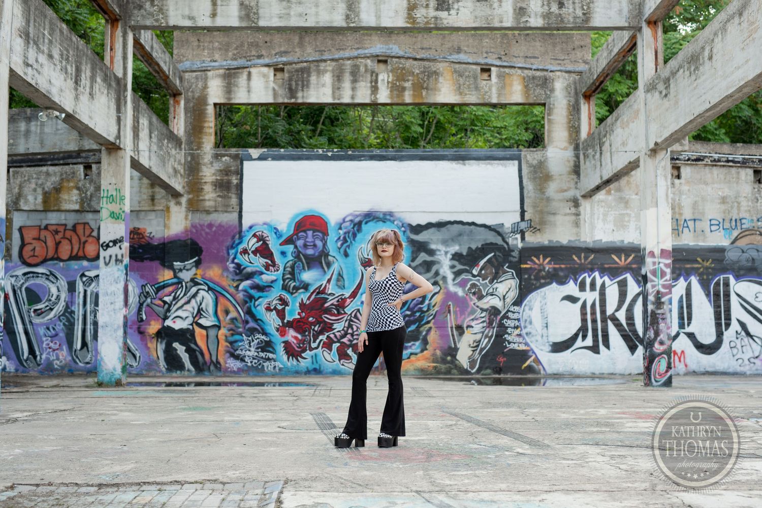 A person in grunge style poses against a wall covered in vibrant street art and graffiti murals.