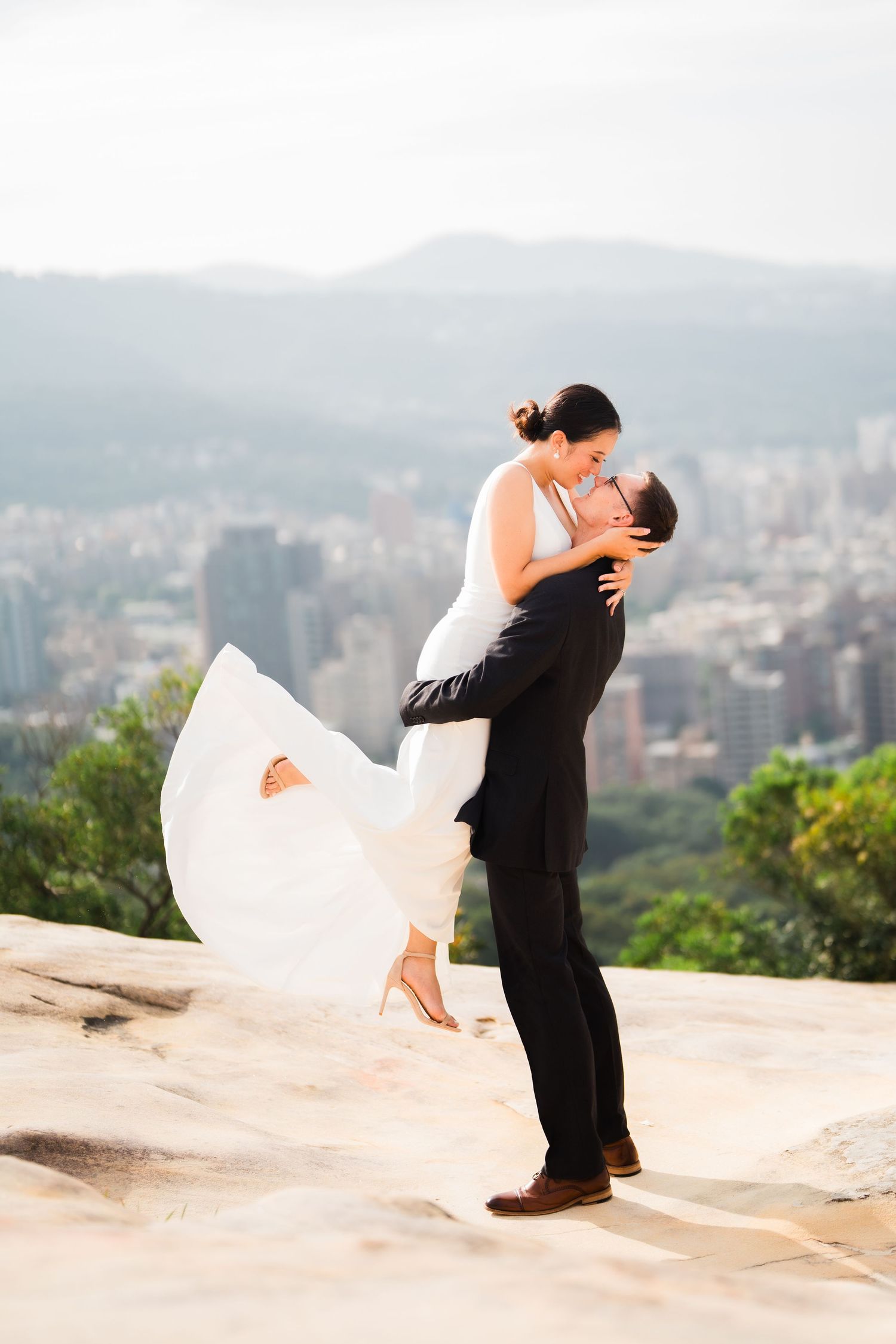 A romantic embrace on a clifftop overlook with a cityscape visible in the background.