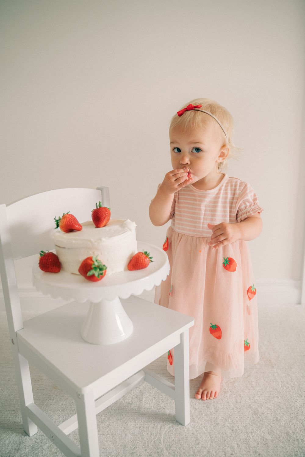 White cake stand with fresh strawberries next to chair against neutral background.