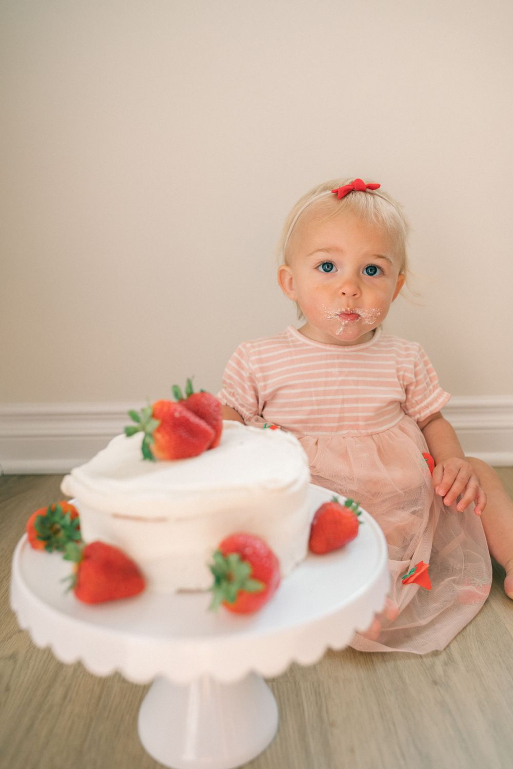 A white cake decorated with fresh strawberries sits on a scalloped pedestal stand on a wooden floor.