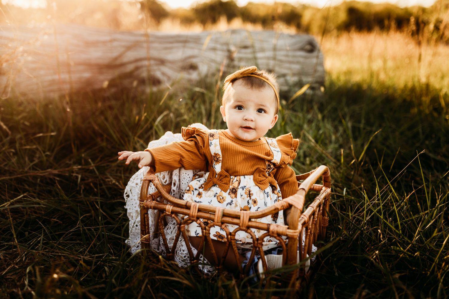 Adorable baby girl in rust orange dress sitting in wicker basket outdoors in golden sunset light.