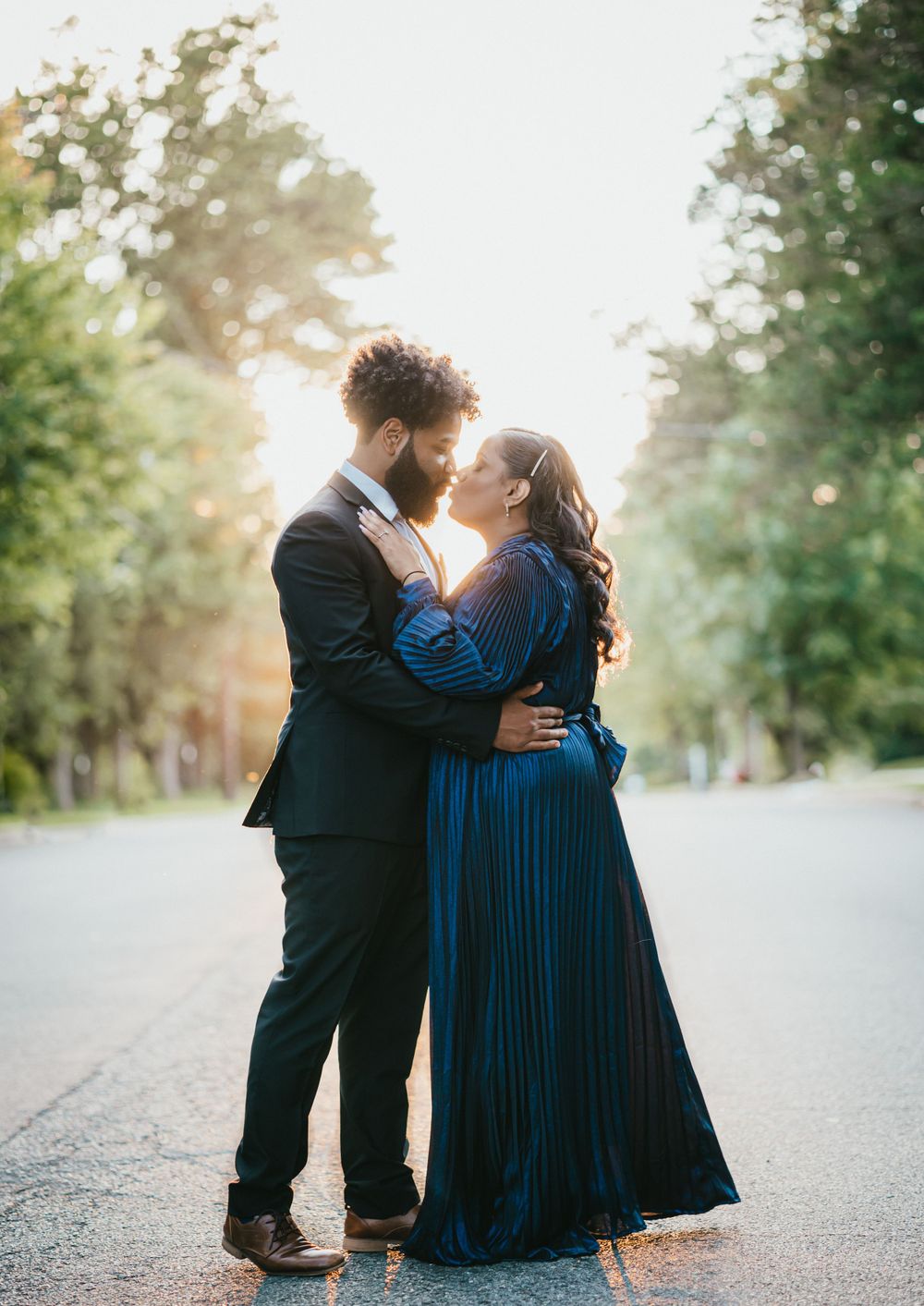 Intimate couple embracing on a tree-lined path at sunset with a flowing blue dress.
