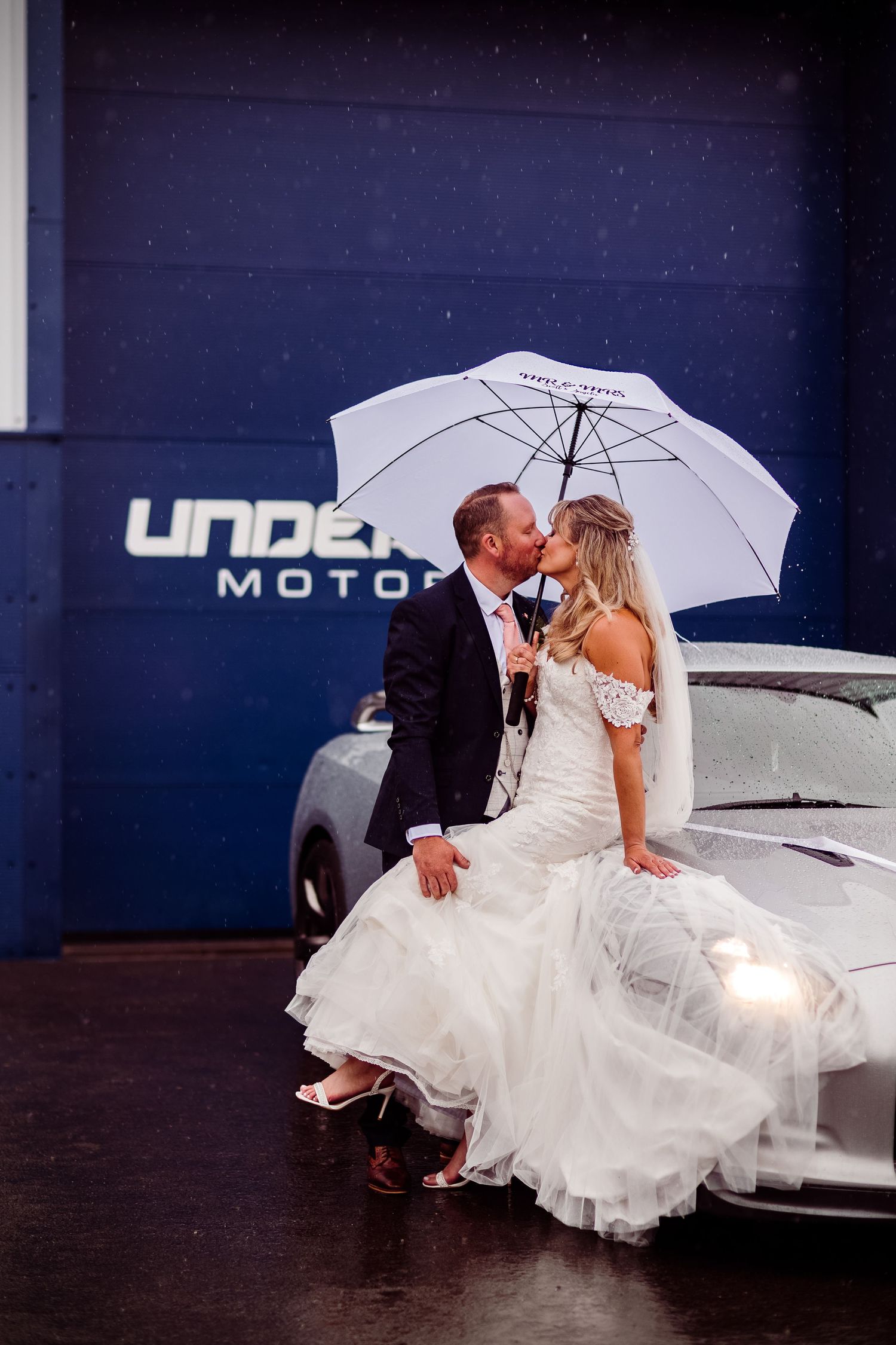 Romantic moment captured with couple posing on sports car under white umbrella.