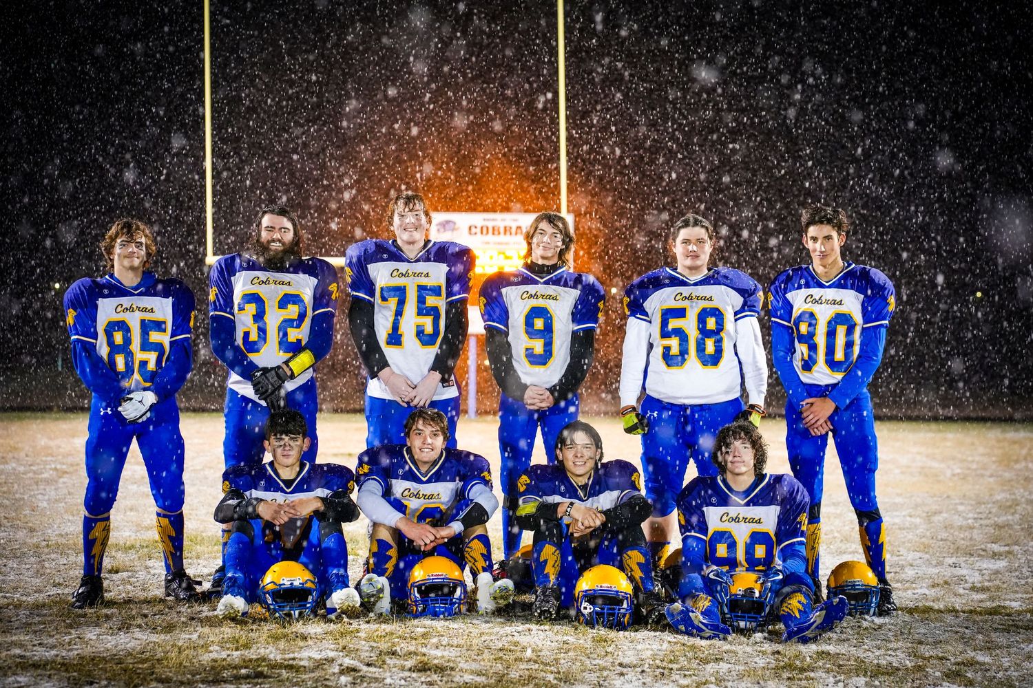 Football team posed in blue uniforms during snowfall at night with stadium lights glowing behind.