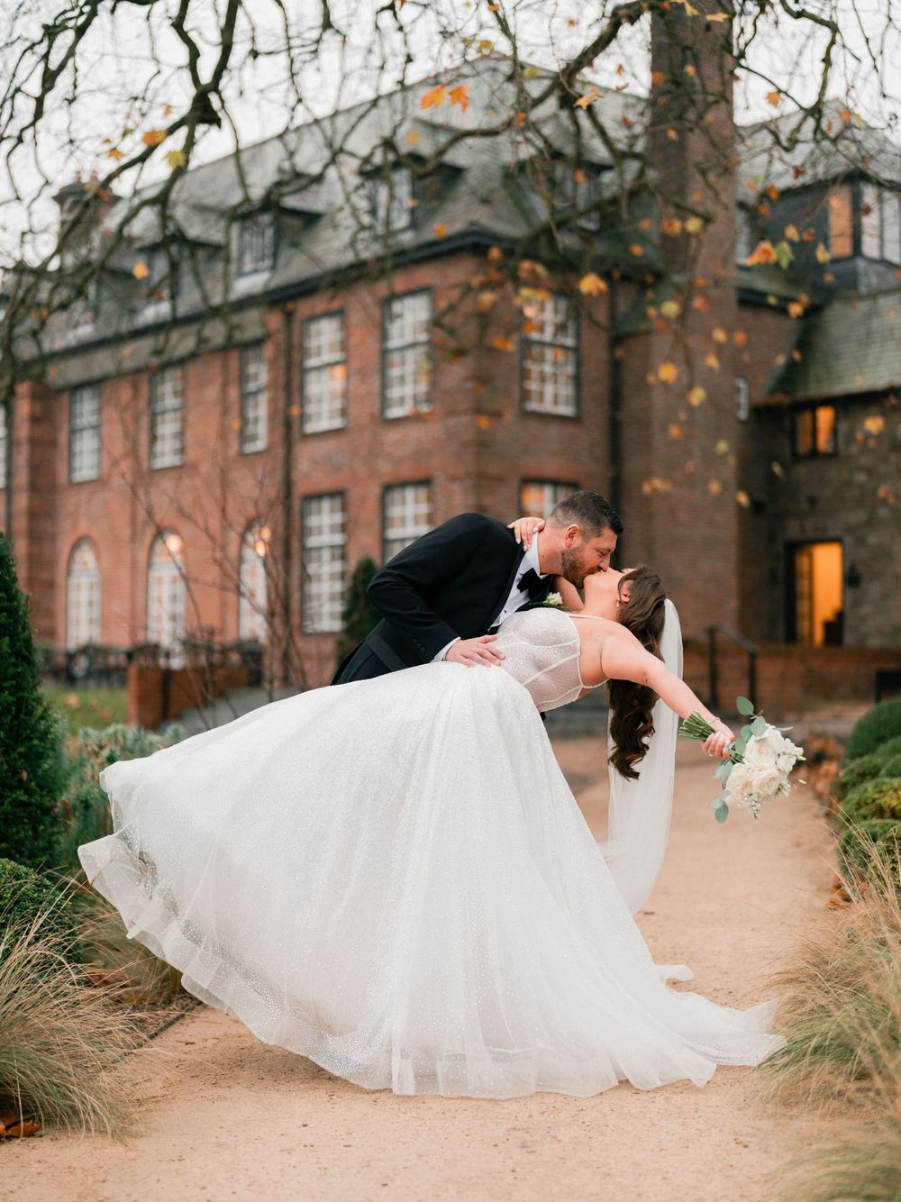 Romantic wedding couple dips and kisses in front of historic brick mansion with fall leaves.