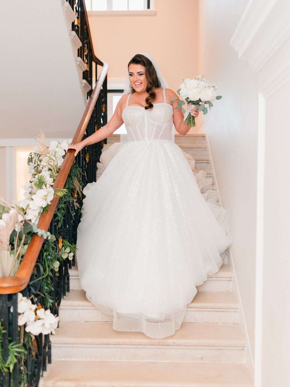 Bride in flowing white ball gown poses elegantly on staircase decorated with floral arrangements.