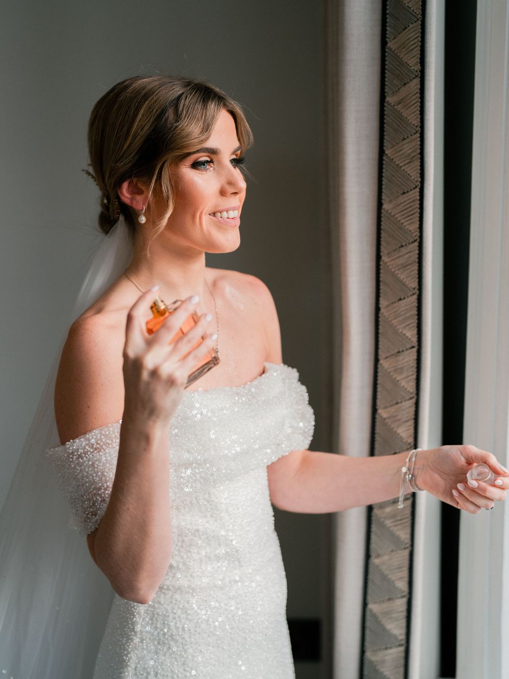 Bride in sparkly white dress poses playfully against gray wall.