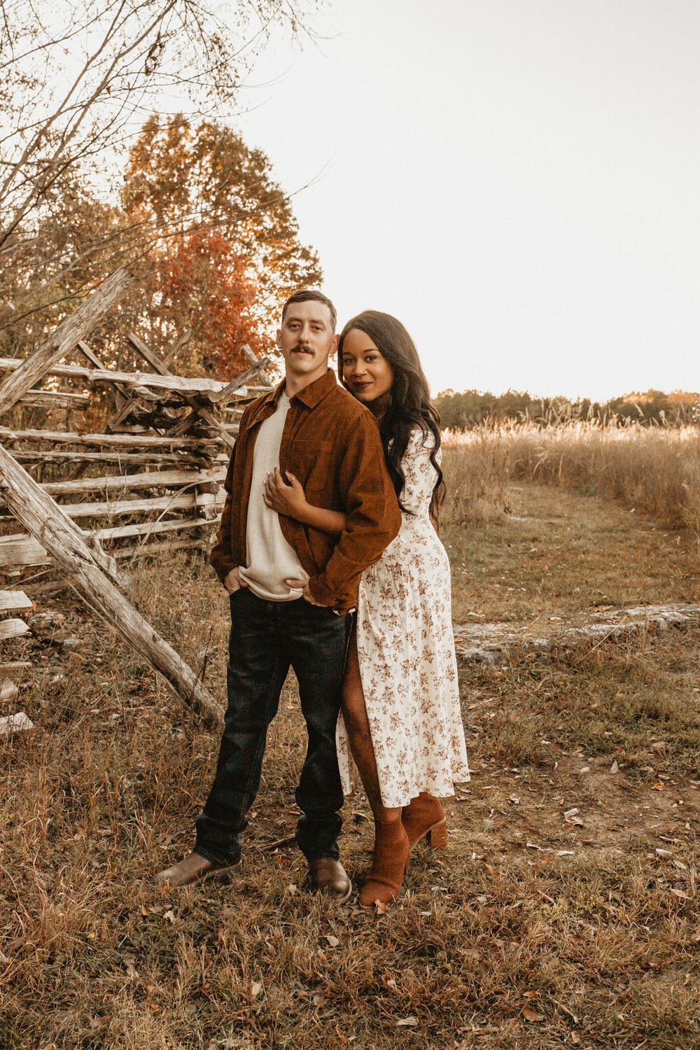 A romantic couple poses together by a rustic wooden fence during autumn golden hour.