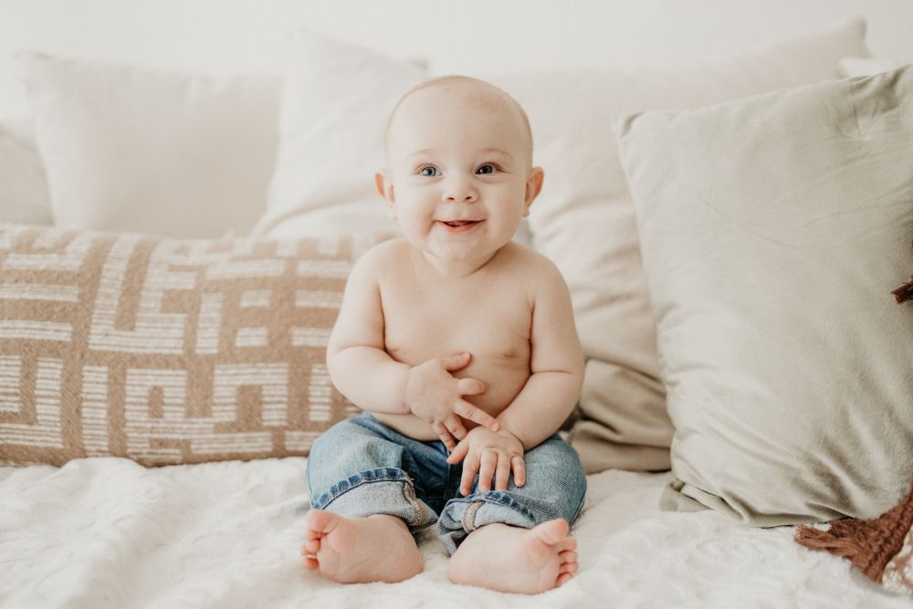 A happy baby in denim shorts sits on white bedding with pillows behind them.