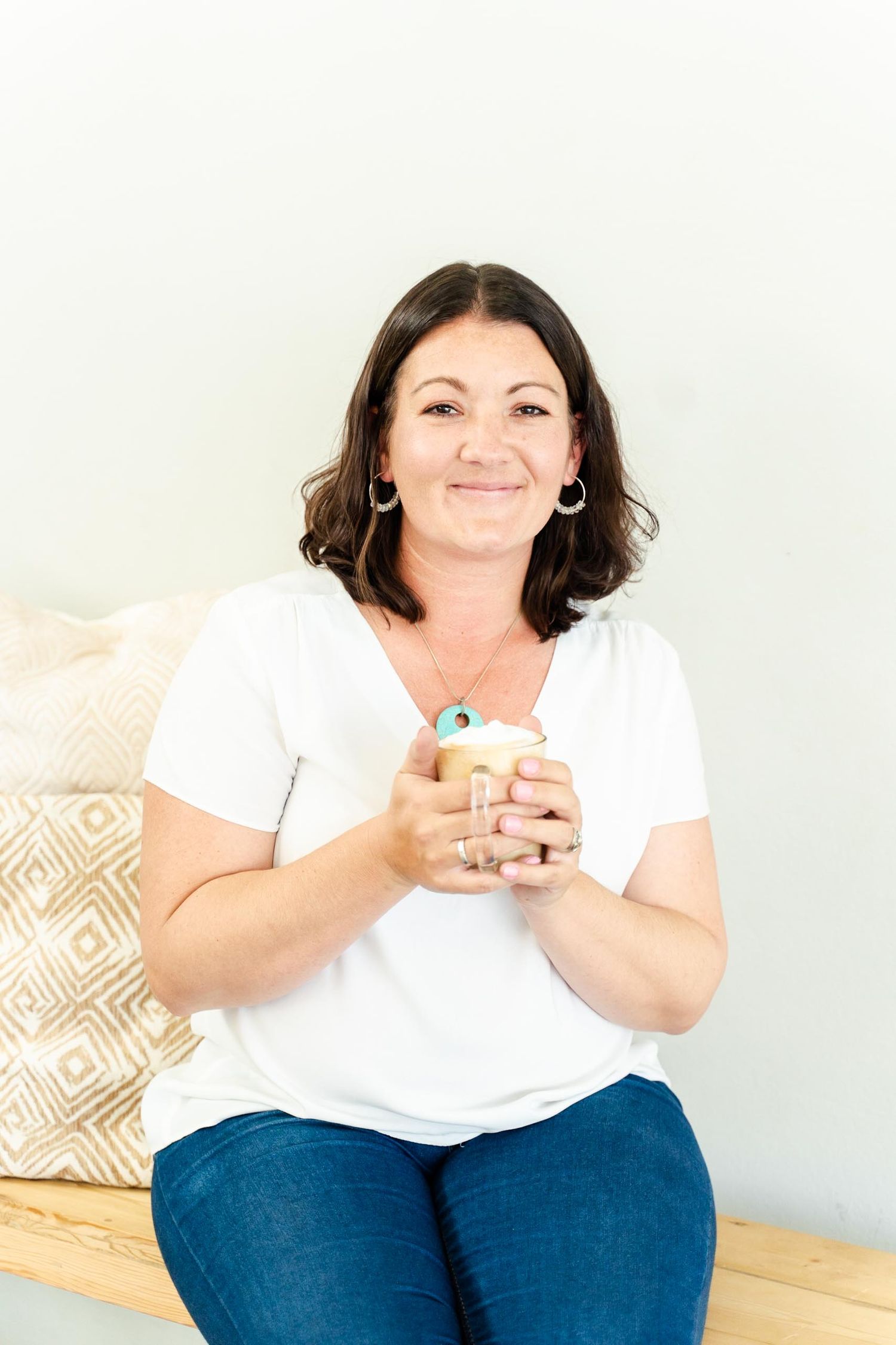 Woman in white t-shirt sits comfortably on bench holding coffee mug against neutral background.