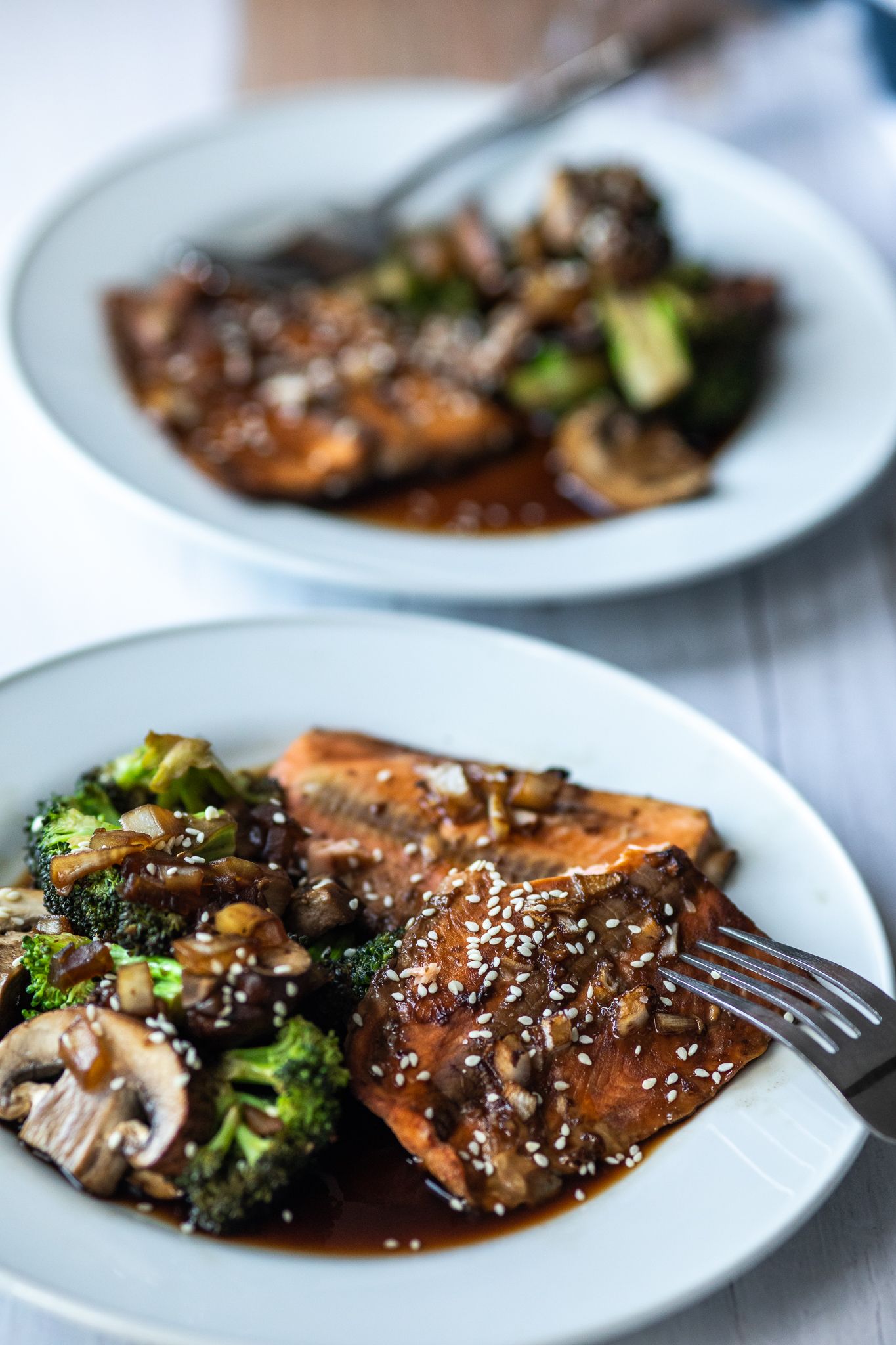 Glazed salmon fillets with broccoli and sesame seeds served on white plates in a teriyaki-style presentation.