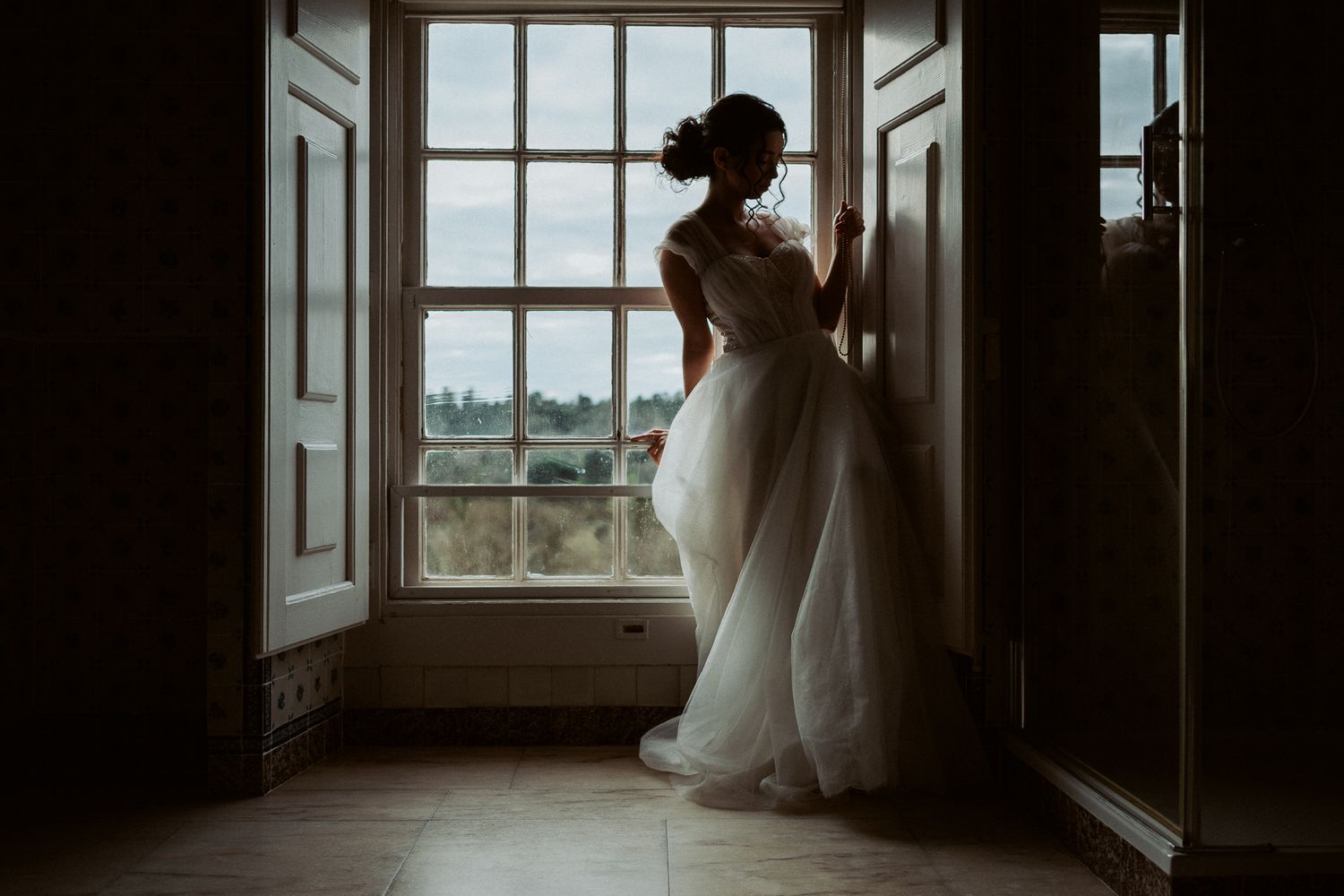 A silhouette stands in an elegant white wedding dress against a bright window in a darkened room.