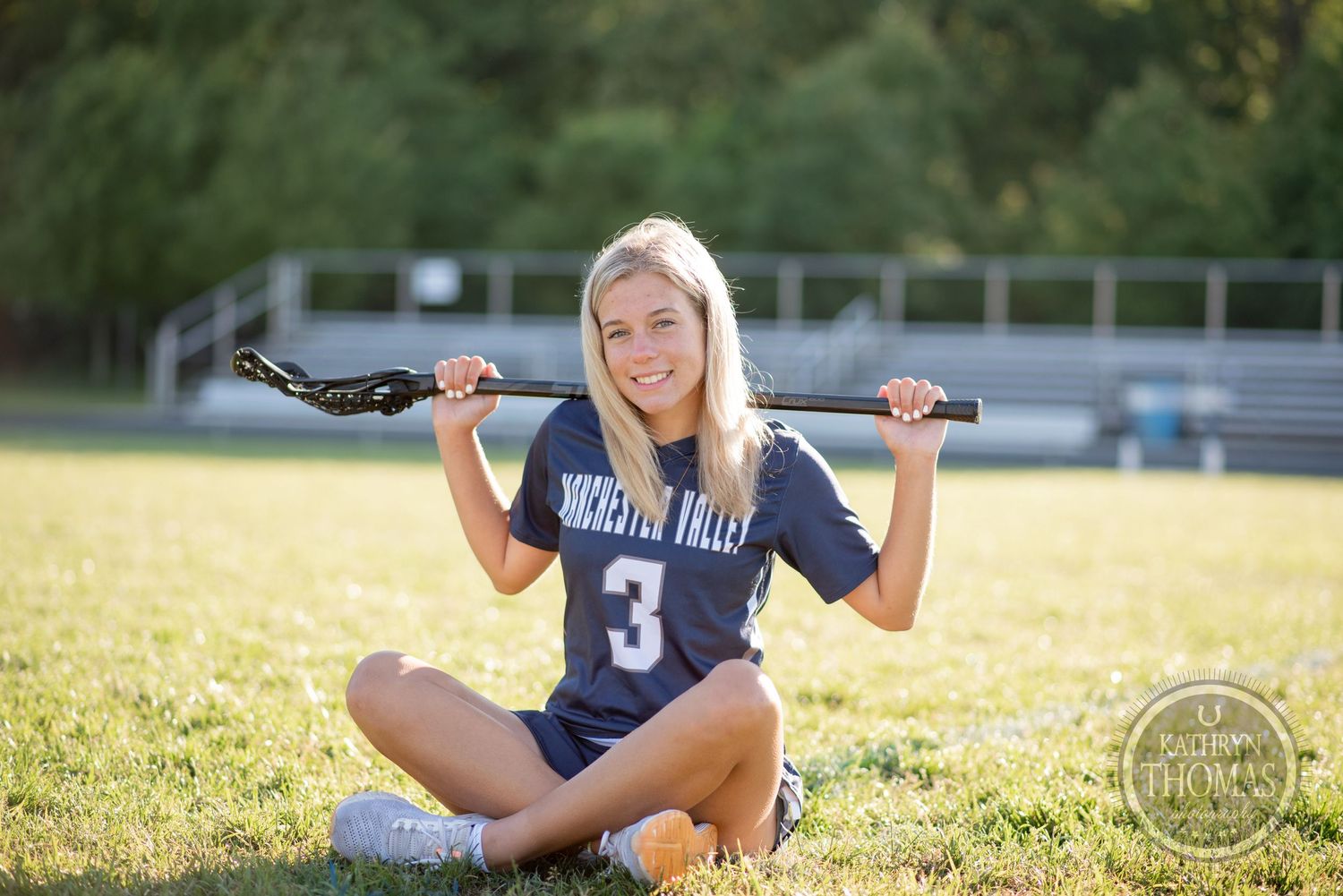 Athlete in navy uniform poses with lacrosse stick on sunny sports field.