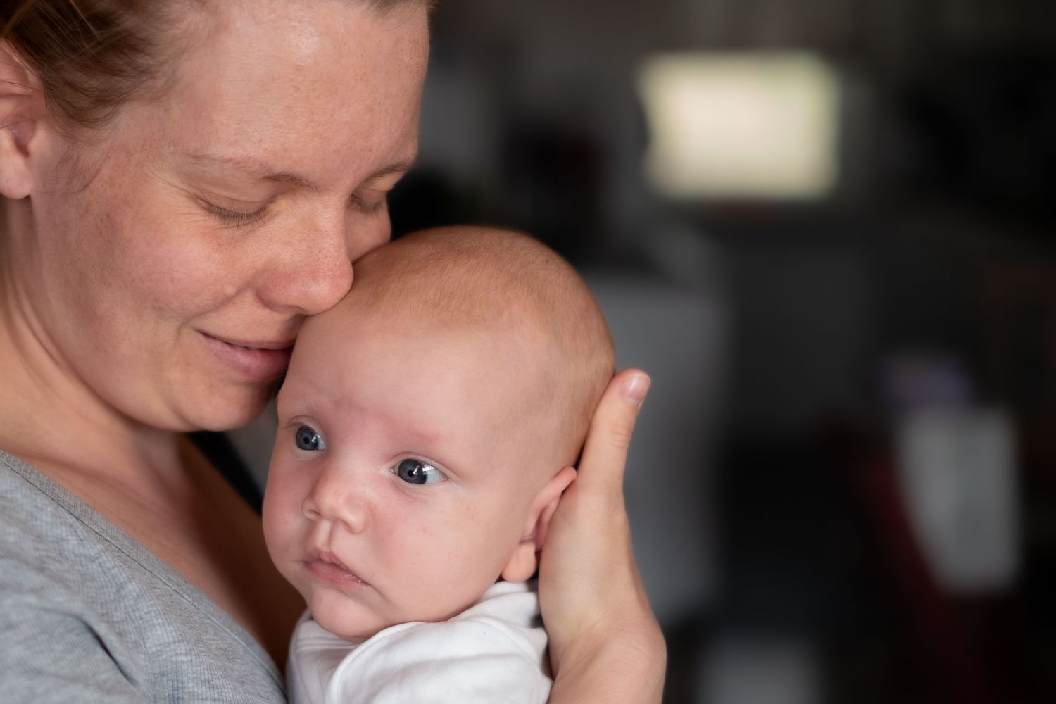 A mother in a gray shirt lovingly cradles and embraces her young baby in a tender intimate moment.