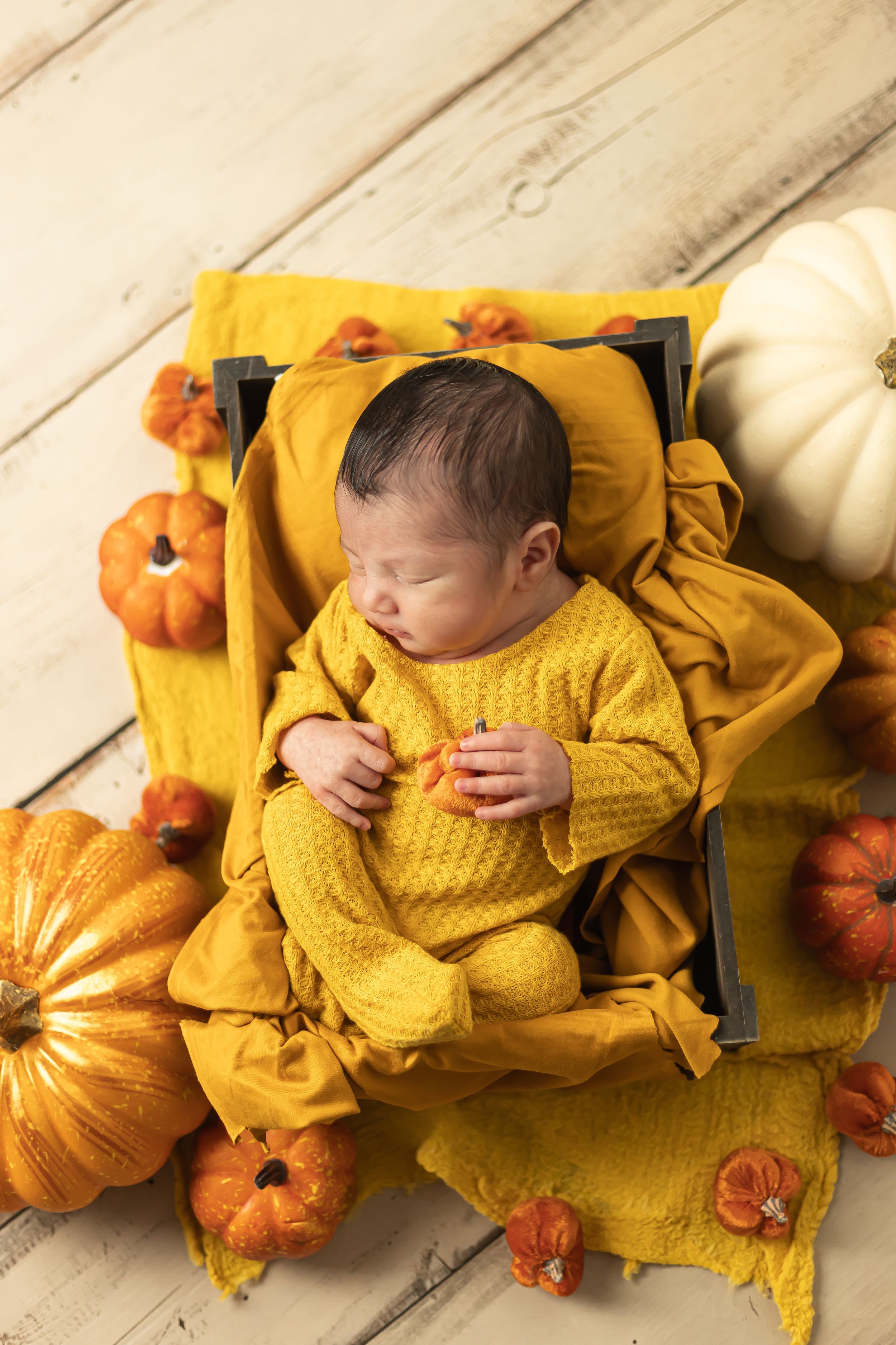 Baby in yellow outfit sleeping among autumn pumpkins on wooden floor.