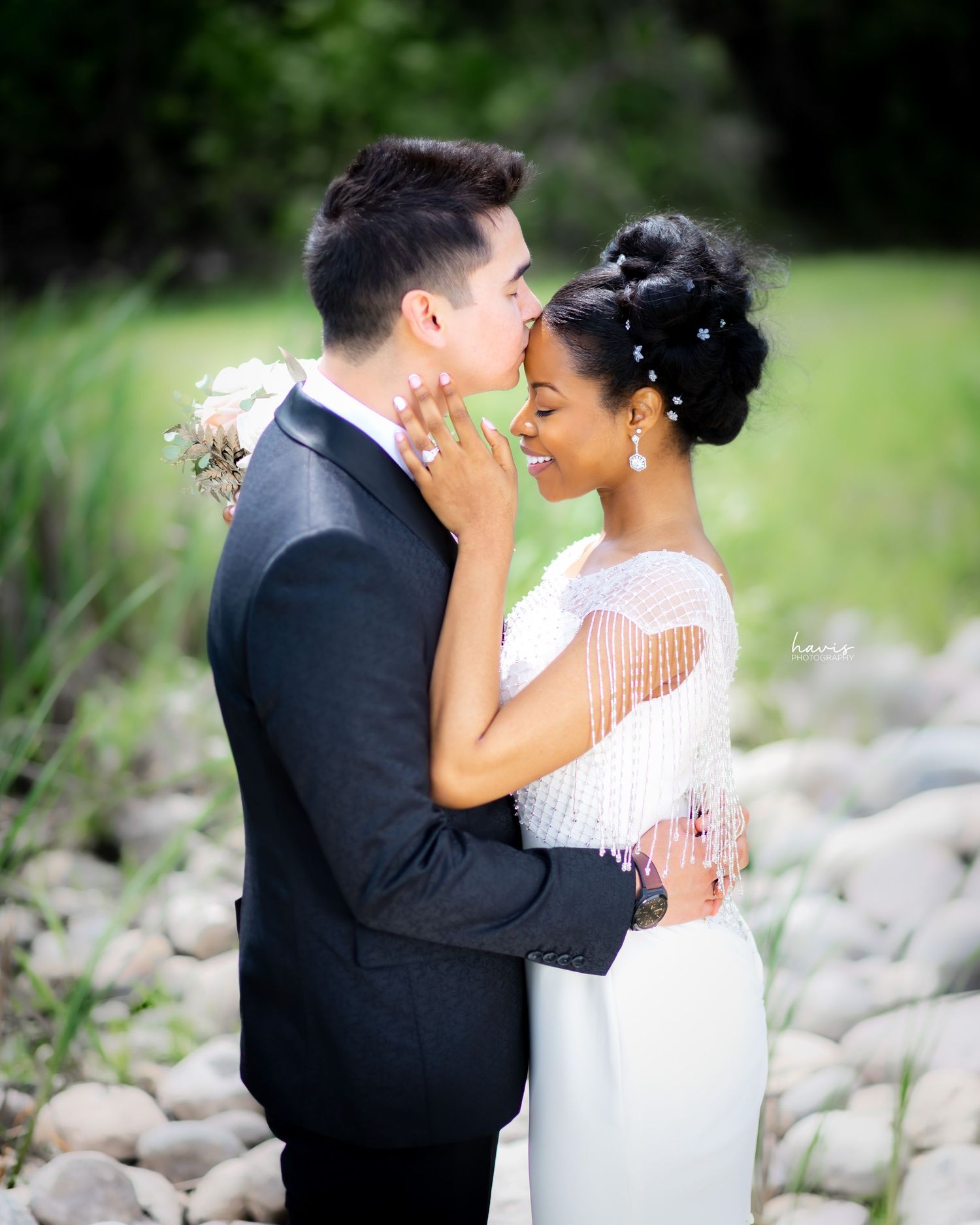 A husband kissing his newly wedded wife on the forehead as they pose for a photograph in a scenic location