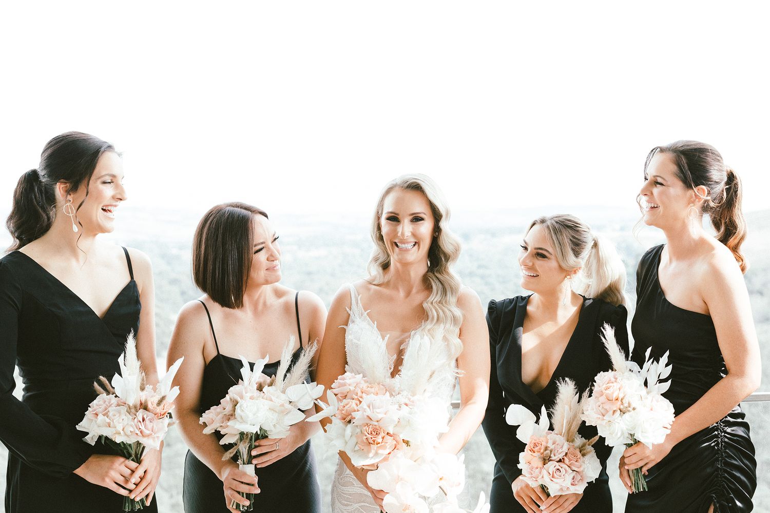 Candid moment of bridal party in black dresses holding pink and white bouquets.