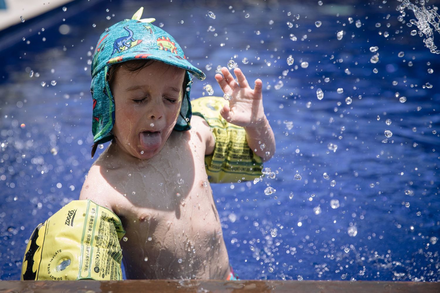 A young swimmer in a blue hat plays in splashing water on a sunny summer day.