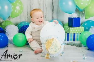 Baby smiling among blue and green balloons with birthday presents in background.