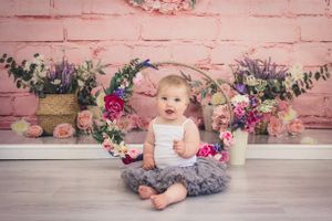 Baby sitting on floor with floral backdrop and rustic pink brick wall.