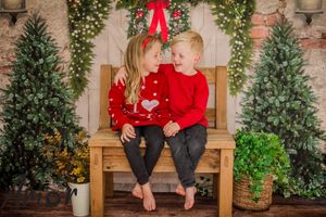 Two children in red holiday sweaters sitting on wooden bench with Christmas decorations in background.