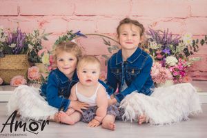 Three siblings in coordinated outfits posing against pink brick wall with floral arrangements.